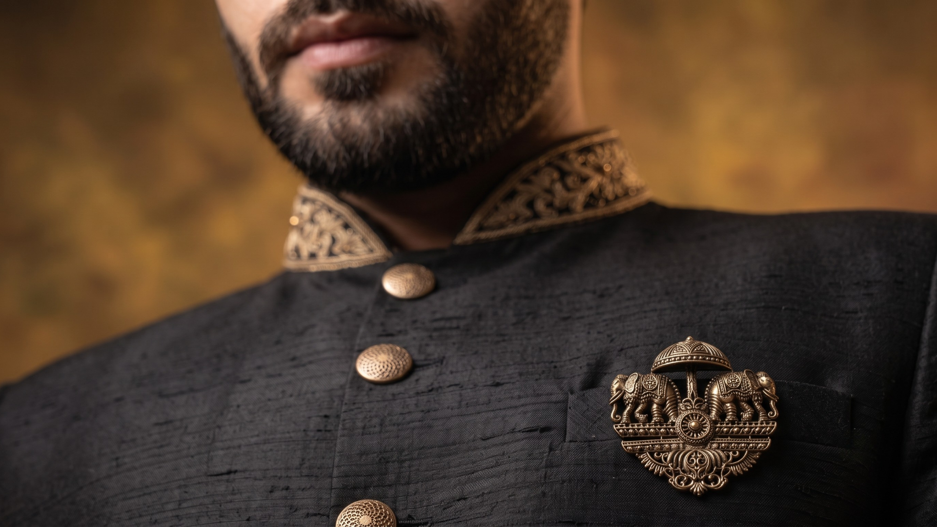 Close-up of a man wearing a black textured sherwani jacket with gold buttons, a patterned gold collar, and a gold elephant emblem brooch on the chest.